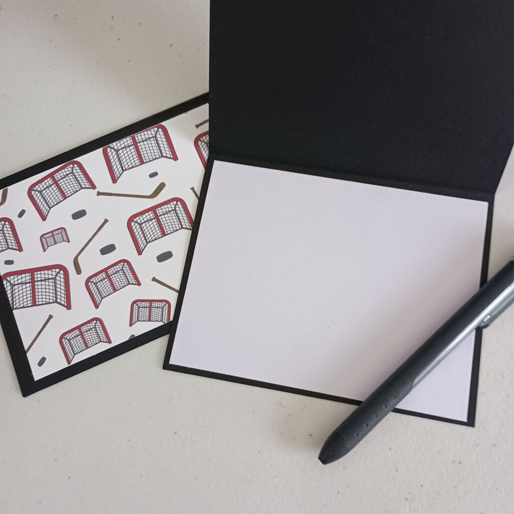 Greeting card with red and black hockey nets, pucks and sticks with an open card with pen showing writing area for personalized message